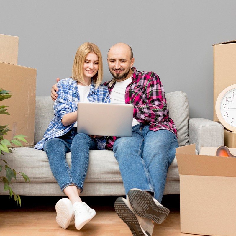 A couple using a laptop on a sofa surrounded by moving boxes